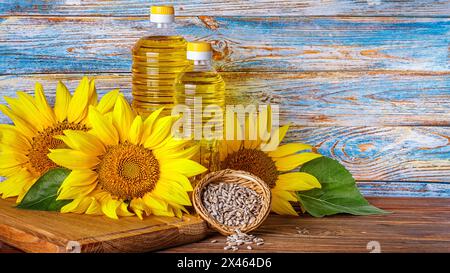 Ländliches Stillleben - Sonnenblumenöl in Flaschen mit Sonnenblumenblüten (Helianthus annuus) und geschälten Samen vor dem Hintergrund einer Holzwand, cl Stockfoto