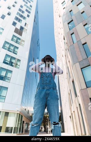 Von unten junger afroamerikanischer Rüde in coolem Outfit Stehen auf der Straße, während Sie Songs in Kopfhörern und hören Blick auf die Kamera Stockfoto