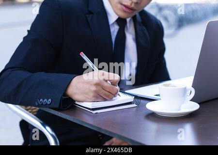Fokussierte asiatische Geschäftsmann in Ohrstöpsel Notizen in Notizblock während Sitzen am Tisch im Café mit Laptop und Planung Geschäft Projekt Stockfoto