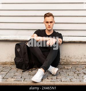 Junger stilvoller Mann in schwarzem Hemd und weißen Schuhen mit Rucksack sitzt in der Nähe der Holzwand Stockfoto