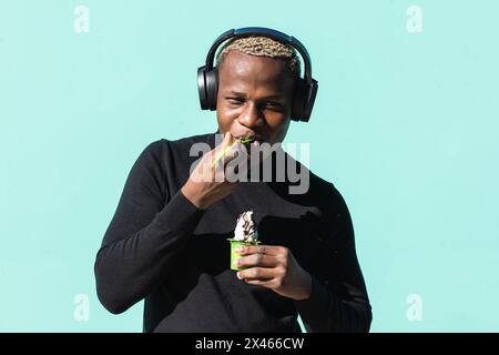 Inhalt junger afroamerikanischer Mann Millennial in lässiger Kleidung zuhören Zu Musik im Kopfhörer und essen leckeres Eis gegen Blauer Hintergrund o Stockfoto
