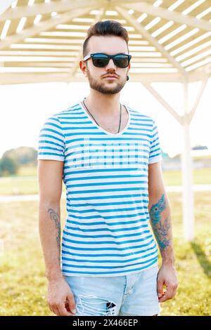 Trendiger schöner Hipster-Mann mit Bart in Sonnenbrille in Einem trendigen Hemd steht am Strand. Stockfoto