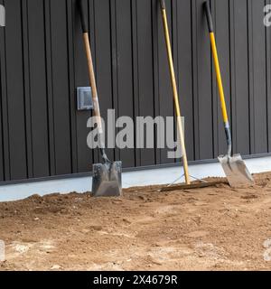 Schmutzige, gebrauchte Schaufeln und ein Rechen stehen aufrecht auf dem Schmutz und lehnen sich an das Brett und die Stanzverkleidung einer Mauer auf einer Baustelle. Stockfoto
