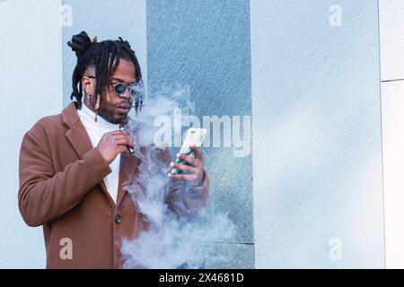 Unemotionaler ernster bärtiger afroamerikanischer Hipster-Mann in stilvollem Outfit Und Sonnenbrillen halten elektronische Zigarette beim Surfen auf dem Smartphone n Stockfoto