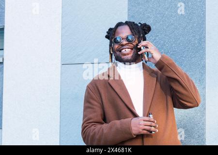 Glücklich bärtig afroamerikanischen Hipster männlich in stilvollem Outfit und Sonnenbrillen halten elektronische Zigarette und lachen während Telefongespräch in der Nähe Stockfoto