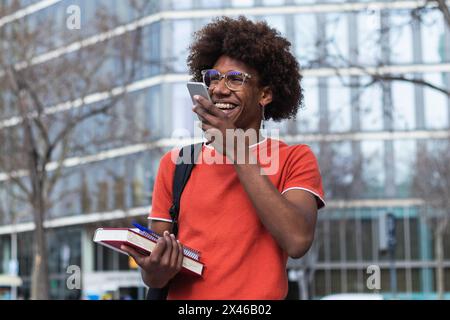 Fröhlicher junger afroamerikanischer Student mit lockigen Haaren Legere Kleidung und Brillen lächeln, während die Sprachnachricht eingeschaltet wird Smartphone in der Stadt Stockfoto