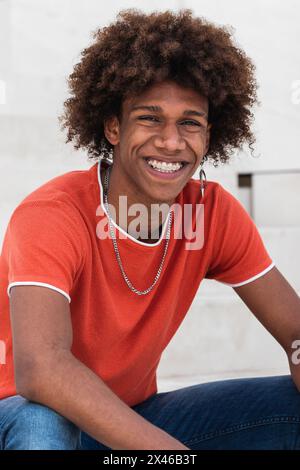 Selbstbewusster junger afroamerikanischer Mann mit lockigen Haaren in stilvoller Form Outfit lächelnd und Blick auf die Kamera, während Sie sich in der Stadt ausruhen Straße Stockfoto