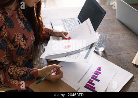 Crop fokussierte Frau in stilvollen Kleid Büroangestellte Analyse von Diagrammen Beim Sitzen am Schreibtisch mit Laptop und Papieren Stockfoto