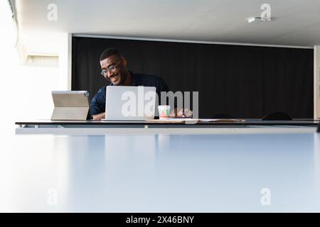 Positiver afroamerikanischer Erwachsener, männlich in formeller Kleidung und Brillen, die am Tisch sitzen, während er einen Videoanruf auf dem Tablet in der Nähe des Netbooks führt und Wi dokumentiert Stockfoto