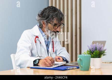 Älterer männlicher Arzt sitzt am Tisch in der Arztpraxis und nimmt sich Notizen, während er in der Klinik arbeitet Stockfoto