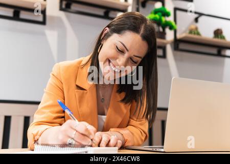 Niedriger Winkel der entzückten weiblichen freiberuflichen Schreiben in Notizblock, während sie am Tisch mit Laptop im Café sitzt und an Remote-Projekt arbeitet Stockfoto