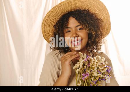 Eine junge afroamerikanische Frau mit lockigen Haaren, die einen Strohhut trägt, hält in einem Atelier einen lebendigen Blumenstrauß. Stockfoto