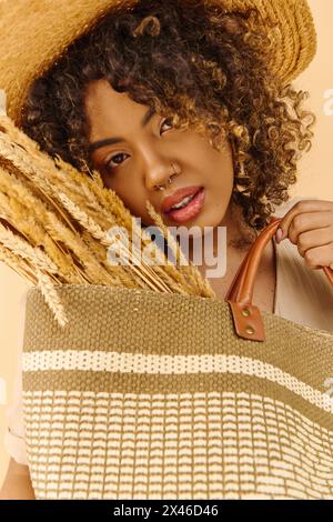 Eine junge Afroamerikanerin mit lockigem Haar, die ein Sommerkleid trägt, eine Tasche hält und einen stylischen Strohhut trägt. Stockfoto