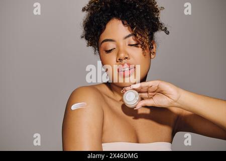 Die junge afroamerikanische Frau mit lockigem Haar trägt wunderbar Creme auf ihr Gesicht in einem Studio-Setting auf. Stockfoto