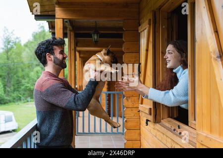 Seitenansicht eines lächelnden Paares von Reisenden, die mit einer entzückenden französischen Bulldogge auf der Terrasse einer Holzhütte in den Pyrenäen spielen Stockfoto