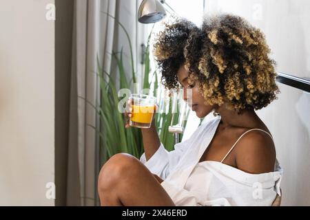Seitenansicht einer afroamerikanischen Frau in weißer Kleidung, die zu Hause frischen Orangensaft aus dem Glas trinkt Stockfoto
