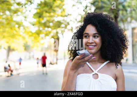 Positive afroamerikanische Frau mit Afro-Frisur, die Sprachnachrichten auf dem Handy auf der Straße der Stadt aufzeichnete Stockfoto