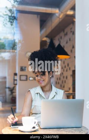 Positive afroamerikanische Freiberuflerin mit grafischem Tablet am Tisch mit Laptop und Kaffee Stockfoto