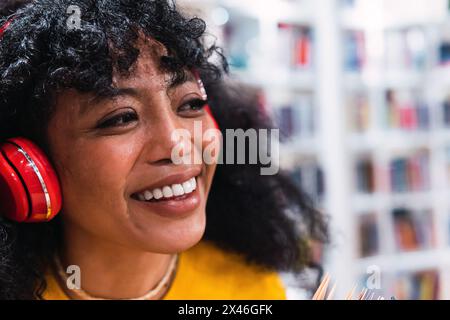 Crop fröhliche junge ethnische Frau mit lockigen dunklen Haaren lächelnd und wegschauen, während Musik in Kopfhörer in der modernen Bibliothek hören Stockfoto