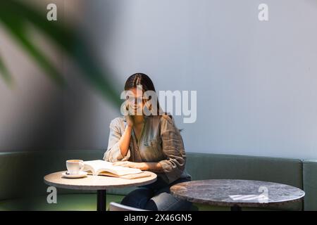 Junge weibliche Inhalte berühren das Gesicht beim Lesen des Lehrbuchs an der Cafeteria mit einer Tasse heißem Getränk Stockfoto