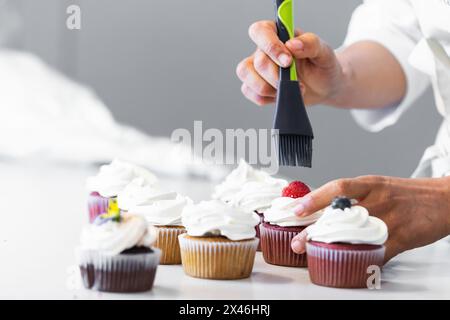 Nicht erkennbarer Fruchtbäcker mit Silikonbürste, der süßen Sirup auf Beeren verteilt, während er in der Küche vegane Cupcakes zubereitet Stockfoto