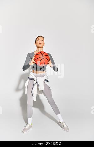 Eine sportliche junge Frau hält elegant einen Basketball in der rechten Hand vor grauem Hintergrund. Stockfoto