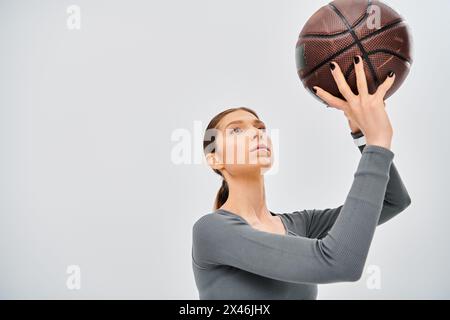 Eine sportliche junge Frau in aktiver Kleidung, die einen Basketball vor grauem Hintergrund in der Luft hält. Stockfoto