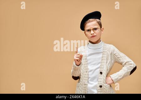Ein vorpubertärer Junge in Brille und Hut posiert als Filmregisseur vor beigefarbenem Hintergrund. Stockfoto
