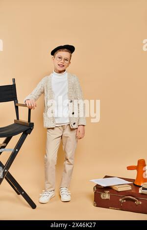 Ein niedlicher Junge, der als Filmregisseur gekleidet ist, steht neben einem Stuhl vor beigefarbenem Hintergrund. Stockfoto