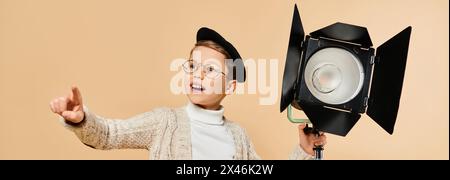 Ein kleiner Junge in Brille und Hut, der eine Kamera hält, verkleidet als Filmregisseur vor beigefarbenem Hintergrund. Stockfoto