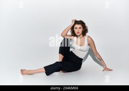 Eine hübsche junge Frau, die anmutig auf dem Boden sitzt, mit überkreuzten Beinen, in schwarzen Hosen und einem weißen Tanktop gekleidet. Stockfoto