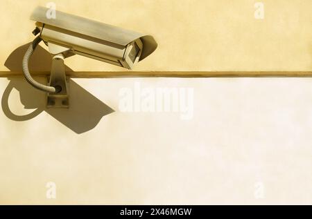 Eine CCTV-Kamera ist an einer beigefarbenen Wand angebracht und überwacht einen offenen Raum mit einem klaren, sonnendurchfluteten Himmel. Stockfoto