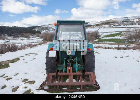 Alter Traktor mit Anbaugerät, das mitten auf einer schneebedeckten Wiese geparkt ist. Berglandwirtschaftskonzept Stockfoto