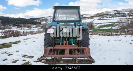 Alter Traktor mit Anbaugerät, das mitten auf einer schneebedeckten Wiese geparkt ist. Berglandwirtschaftskonzept Stockfoto