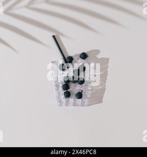 Glas kalter erfrischender Heidelbeerdrink aus Eiswürfeln und Reifen Heidelbeeren mit Palmenblattschatten auf weißem Hintergrund. Kreatives Sommergetränk Stockfoto