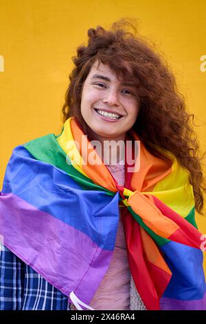 Porträt von fröhlichem, nicht binärem rotem Haar mit Regenbogenfahne über den Schultern, die vor der Kamera lächelt. Stockfoto