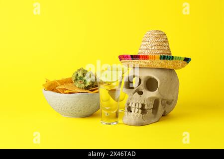 Mexikanischer Sombrero-Hut, menschlicher Schädel, Tequila, Nachos-Chips und Guacamole in der Schüssel auf gelbem Hintergrund Stockfoto