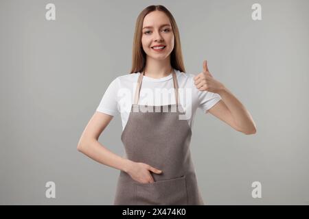 Schöne junge Frau in der Küchenschürze, die Daumen hoch auf grauem Hintergrund zeigt. Modell für Design Stockfoto