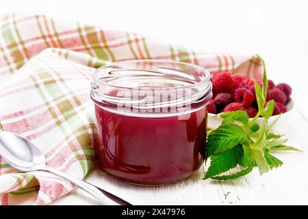 Himbeermarmelade im Glas, Beeren in der Schüssel, grüne Blätter, Serviette und Löffel auf Holzbrett Hintergrund Stockfoto
