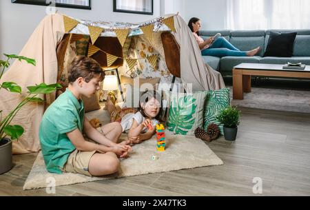 Das kleine Mädchen fängt ein Stück Stapelspiel, während es mit Bruder und Mutter spielt, das Buch auf dem Sofa liest Stockfoto