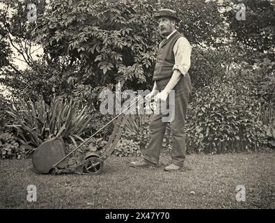 Ein Gärtner, der seinen Rasen im Vereinigten Königreich um 1902 mäht – er verwendet einen Godiva-Handmäher von Barford & Perkins und trägt seine Gartenkleidung. Die Godiva wurde von Barford & Perkins aus Peterborough, Cambridgeshire, England, am Ende des 19. Und Anfang des 20. Jahrhunderts hergestellt. Es enthielt eine Vorrichtung (Wansbrough’s Patent Height Regulator), die 1902 eingeführt wurde. Mit dieser langen Verbindungsstange wurde die Höhe der vorderen Holzwalze eingestellt. Dies wiederum veränderte die Schnitthöhe der Maschine – ein viktorianisches und edwardianisches Foto. Stockfoto