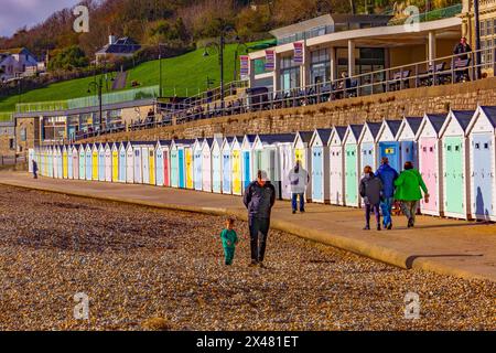 Mann mit einem kleinen Jungen, der am Strand spaziert, während die Wintersonne die farbenfrohen Strandhütten entlang der Küste in Lyme Regis, Dorset, England, erleuchtet Stockfoto