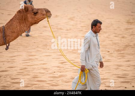 Mann in traditioneller Kurta-Pyjama-Kleidung, der ein Kamel mitten in den Sanddünen führt, auf der Suche nach Touristen auf einer Wüstensafari Stockfoto