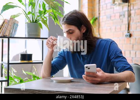 Junger gutaussehender Mann, der Smartphone benutzt, eine Tasse Kaffee trinkt, im Café sitzt Stockfoto