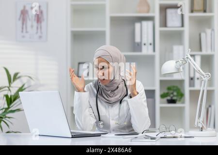 Eine Ärztin, die einen Hijab trägt, zeigt Anzeichen von Frustration und Verwirrung, während sie ihren Laptop in einem hellen medizinischen Büro benutzt. Ihr Ausdruck vermittelt Stress und Schwierigkeiten. Stockfoto