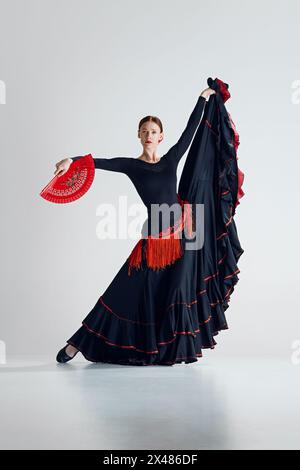 Elegante Frau, Flamenco-Tänzerin mit Fan in der Hand, schwarzes und rotes Kleid, das vor grauem Studio-Hintergrund auftritt Stockfoto