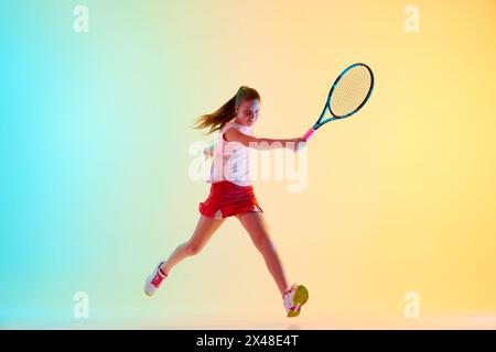 Die junge Tennisspielerin springt in Aktion, ihr Schläger war bereit, einen kräftigen Vorhandschuss in Neonlicht vor blau-gelbem Hintergrund zu treffen. Stockfoto