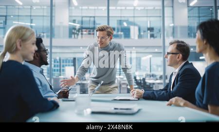 Modern Office Meeting Room: Der hübsche Executive Director steht im Leiter der Konferenz, der sich darauf stützt und eine beredte Rede vor einer Gruppe von Geschäftsleuten und Investoren hält. Stockfoto