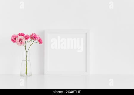 Weißes quadratisches Rahmenmockup mit rosa Rosen in einer Glasvase auf einem weißen Tisch. Stockfoto