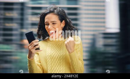Street Shot: Porträt einer schönen lateinischen Frau mit Smartphone, die mit Ja-Geste den erfolgreichen Sieg feiert. Lächelnde Hispanische Unternehmerin Mit Handy Glücklich. Bewegungsunschärfe. Stockfoto